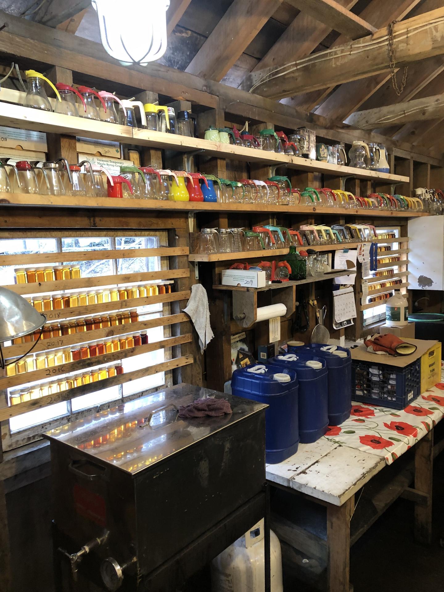 Rustic workshop with shelves full of jars, three blue jugs on a table, and ambient lighting.