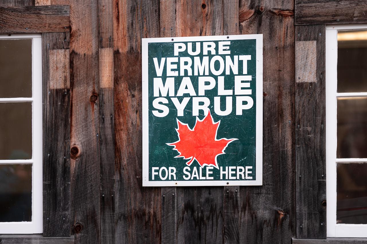 Sign on wooden wall reads "Pure Vermont Maple Syrup" with red maple leaf.