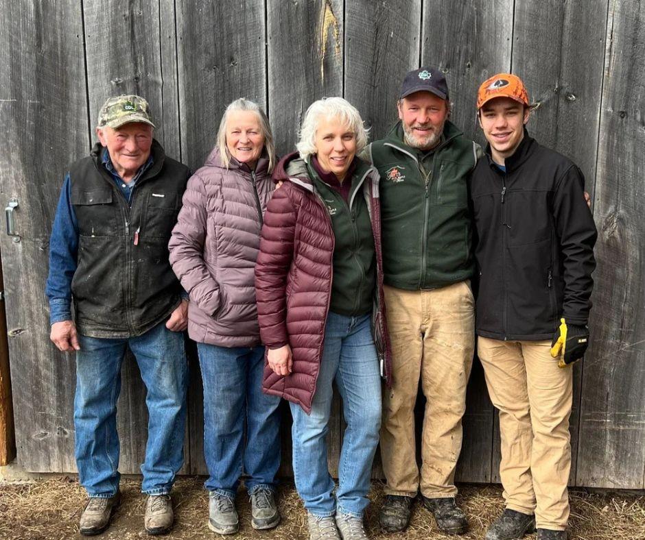 Five people wearing jackets, standing against a wooden wall, smiling at the camera.