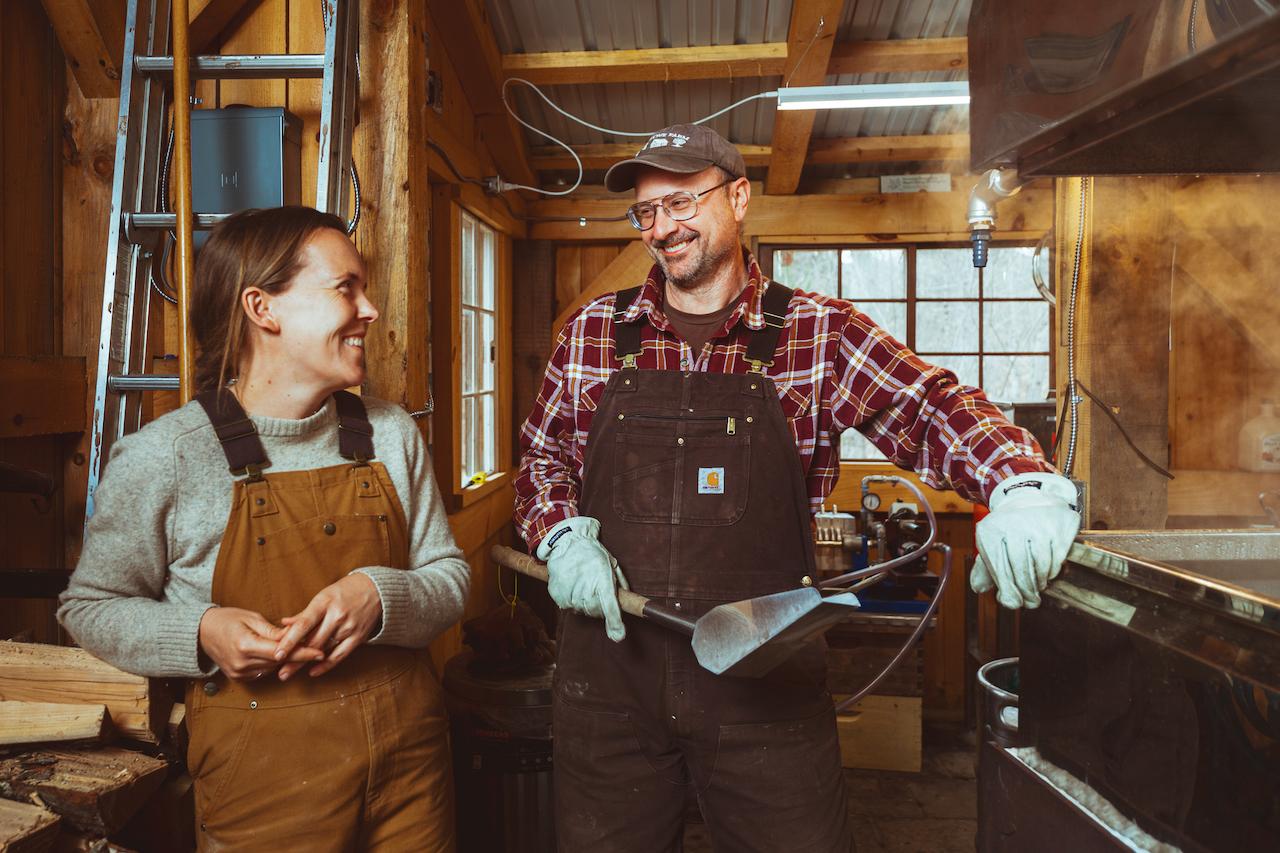Two people in overalls smiling in a cozy, rustic workshop.