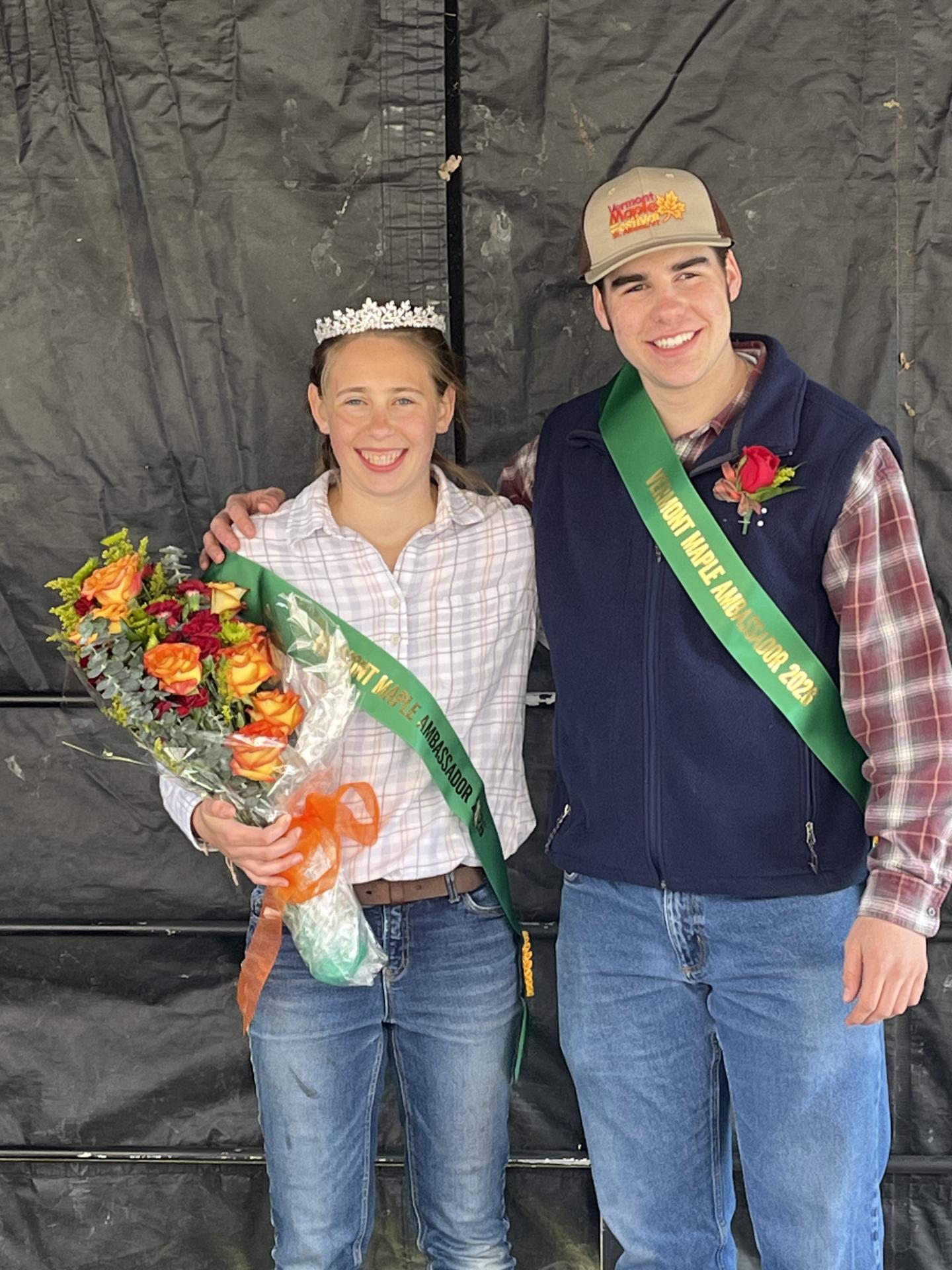 Two people with sashes and bouquet, one wearing a crown and both smiling.