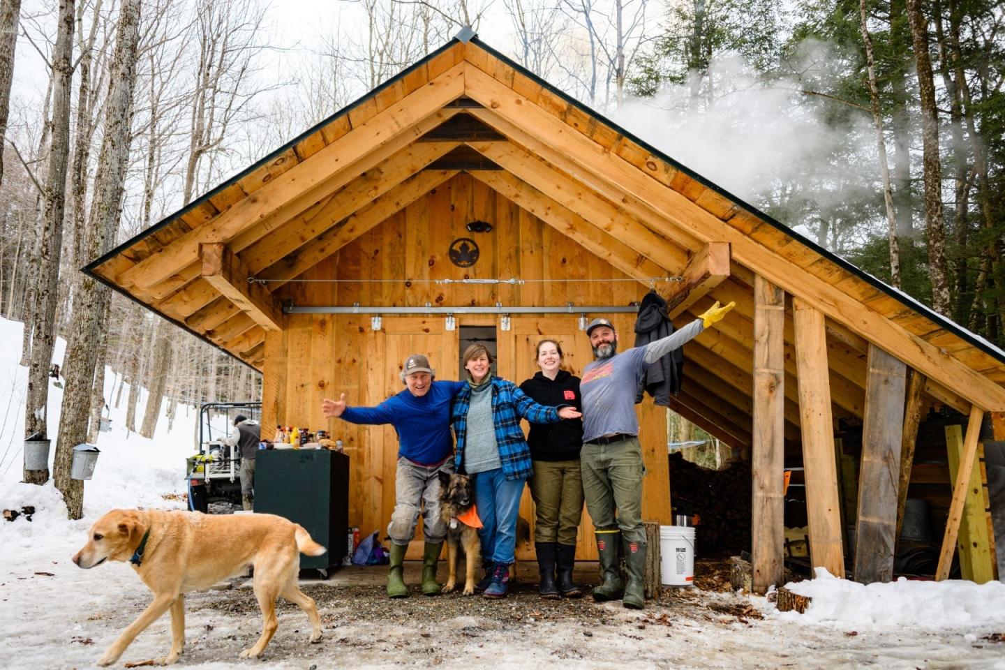 Four people and a dog outside a wooden cabin in a snowy forest.