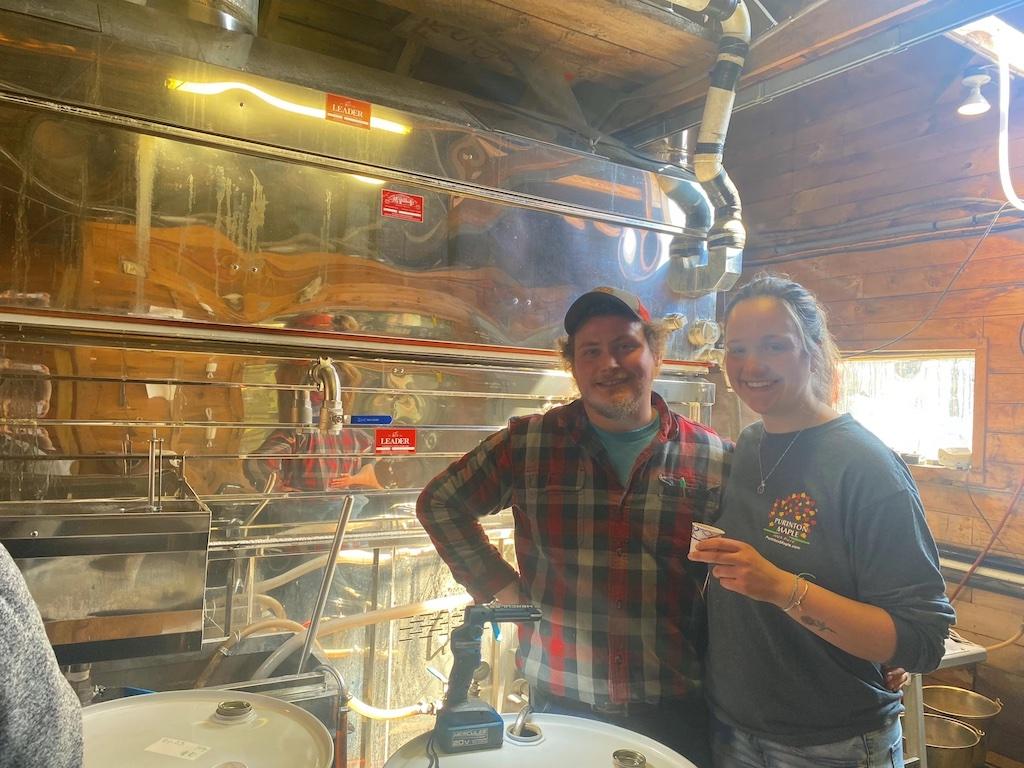 Man and woman smiling in a rustic workshop with metal pipes.