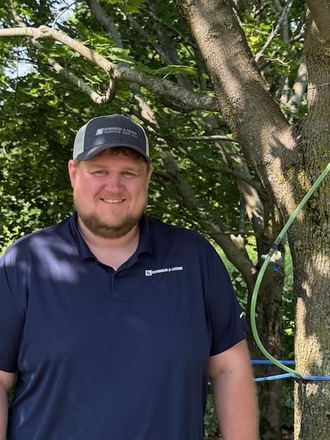 Man in a navy shirt and cap stands by a tree in a sunny setting.