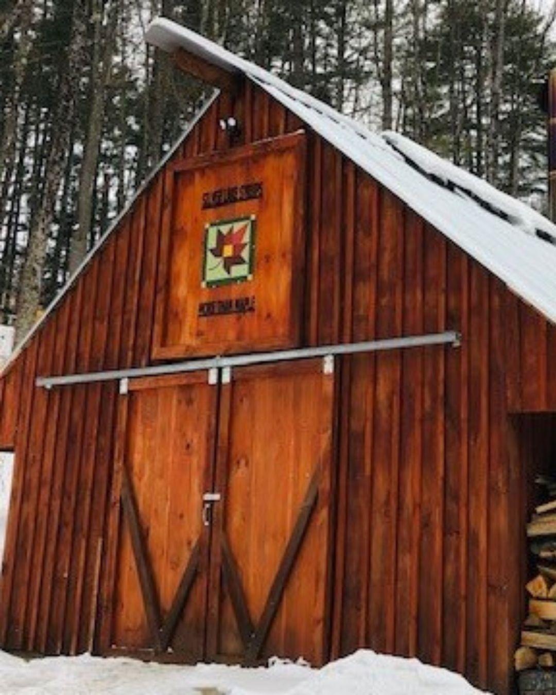 Wooden barn with snow-covered roof in a forest setting.