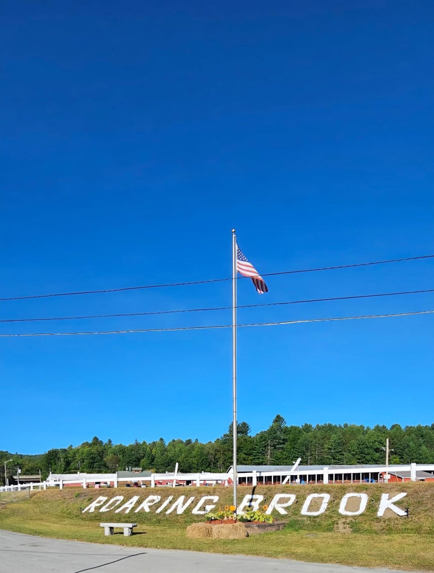 American flagpole against a bright blue sky, with trees and white fence below.