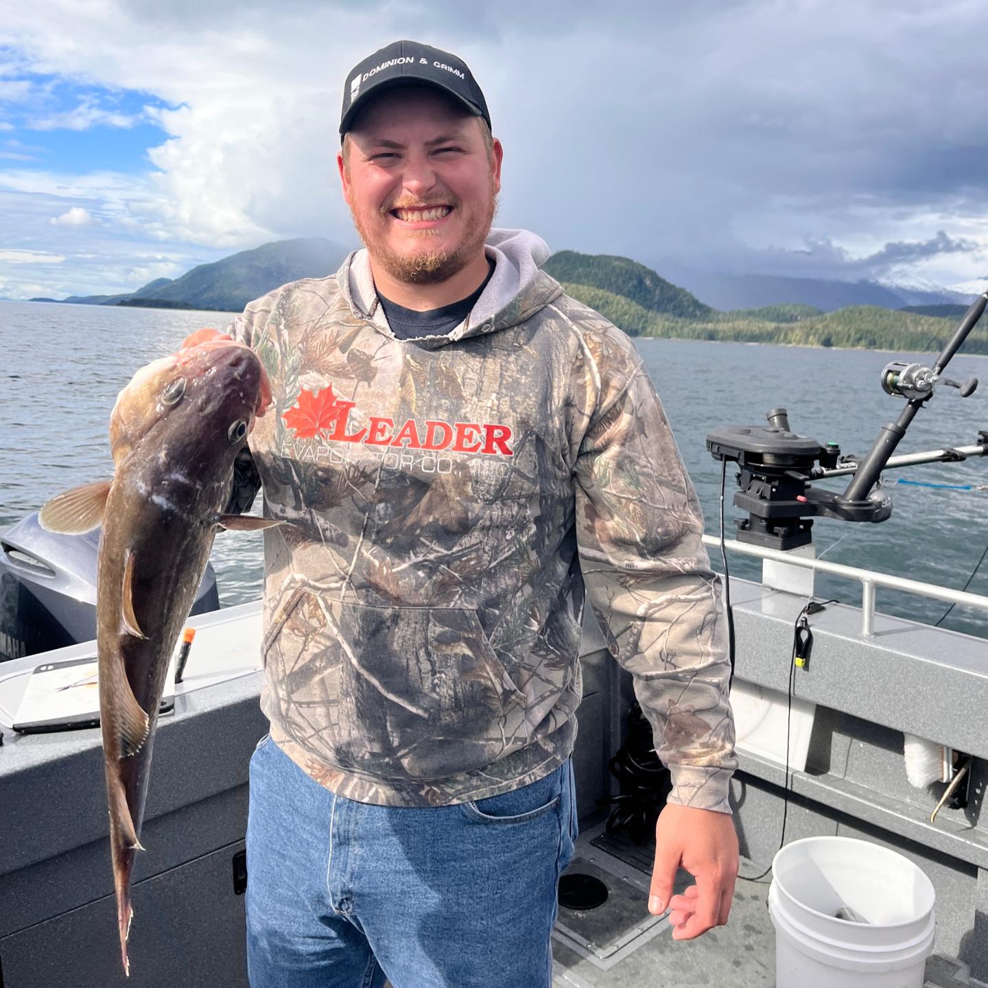 Smiling man holds a fish on a boat, with a lake and mountains in the background.