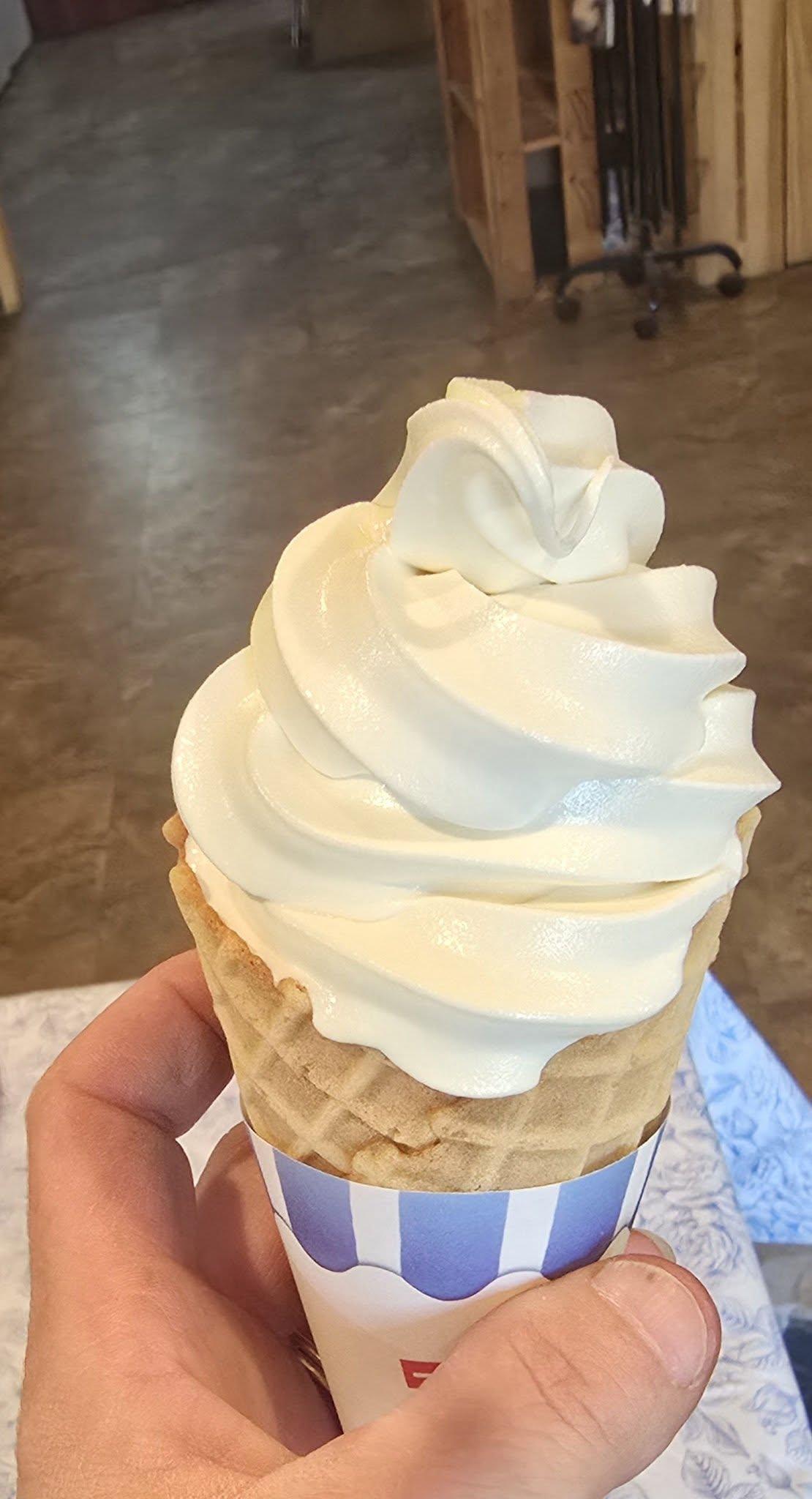 Vanilla soft serve in a waffle cone held by a hand indoors.