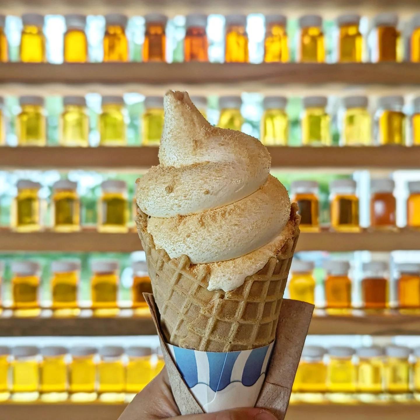 Soft serve ice cream in a waffle cone, held in front of shelves with jars of honey.