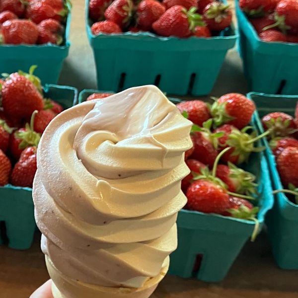 Soft serve ice cream cone held in front of baskets of fresh strawberries.